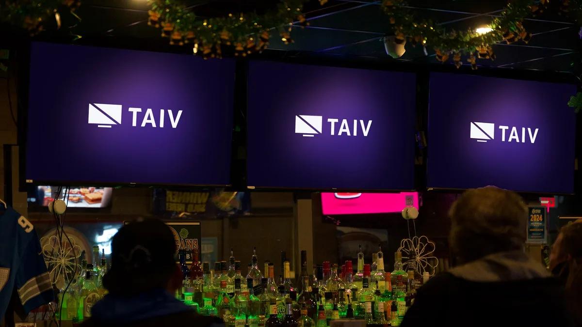 Three Taiv screens mounted above a bar back-bar, with patrons watching in a dim venue
