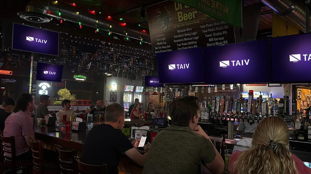 Regulars at a busy neighborhood sports bar with a row of Taiv screens above the taps