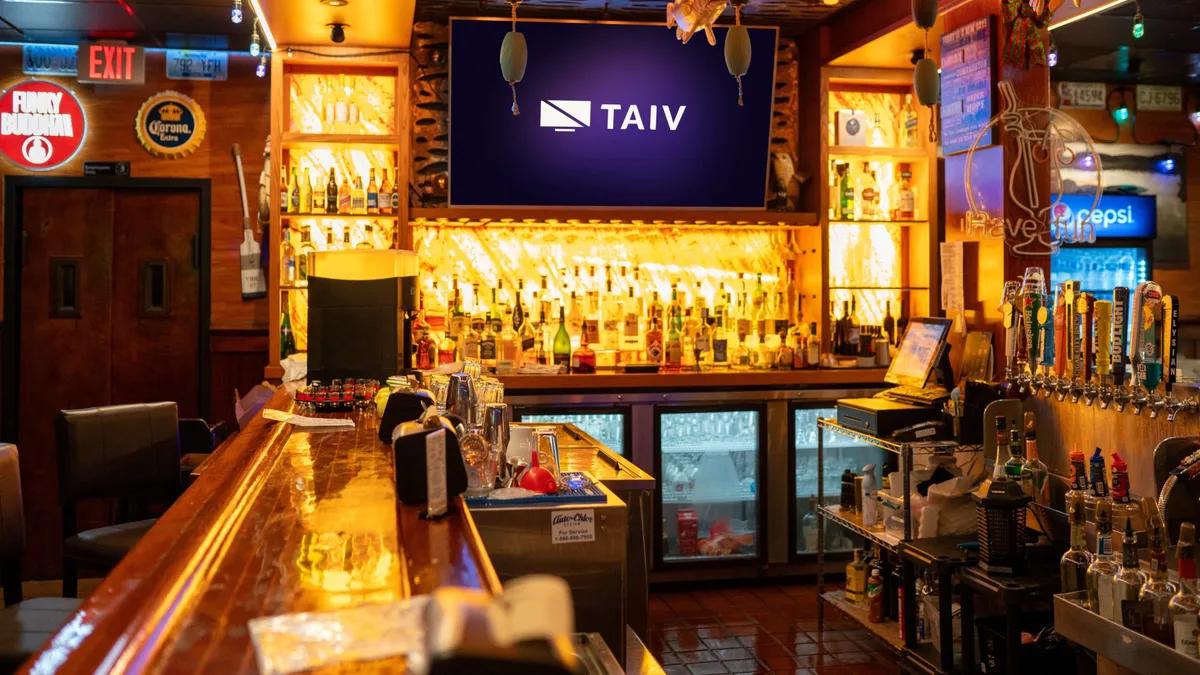 A warm-lit bar with a single Taiv screen mounted above the back-bar and taps