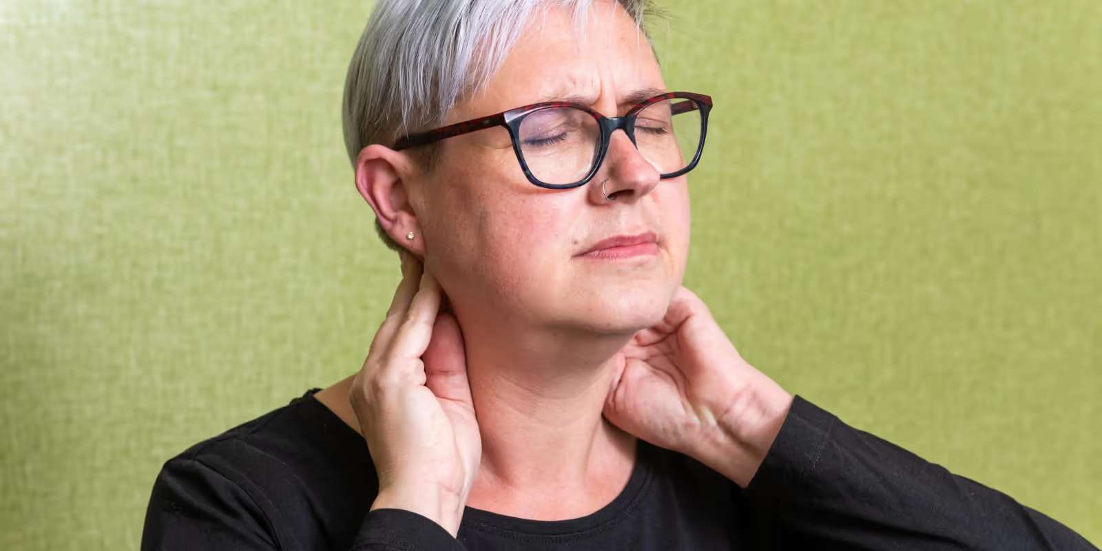 A woman with grey hair and glasses, eyes closed, holding the back of her neck with both hands