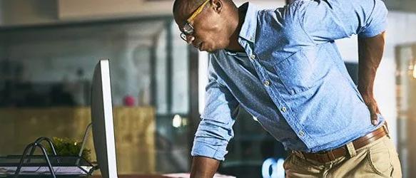 Man in a denim shirt leaning over a desk, holding his lower back