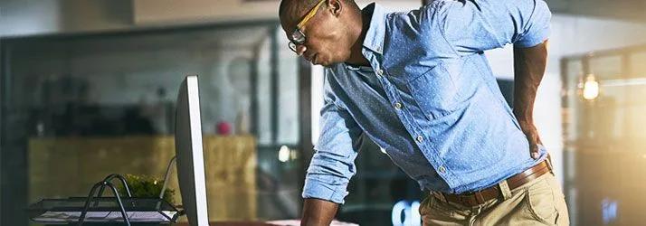 Man in a denim shirt leaning over a desk, holding his lower back