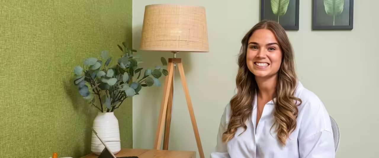 A smiling woman in a white top seated at a desk in a green-walled room with a lamp and a plant