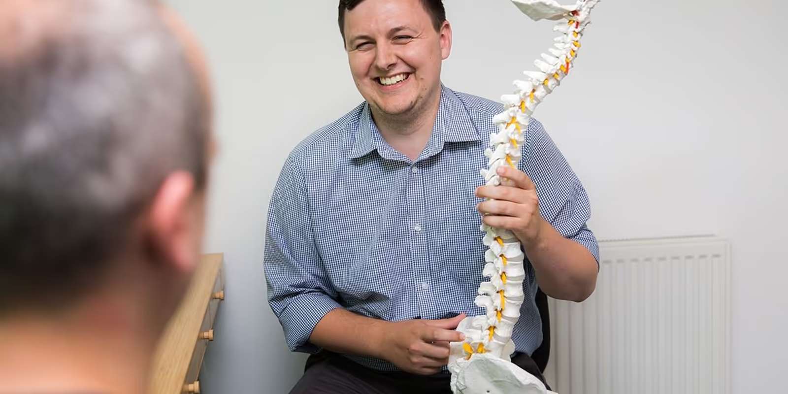 A smiling chiropractor in a checked shirt holding a model of the spine, facing a patient