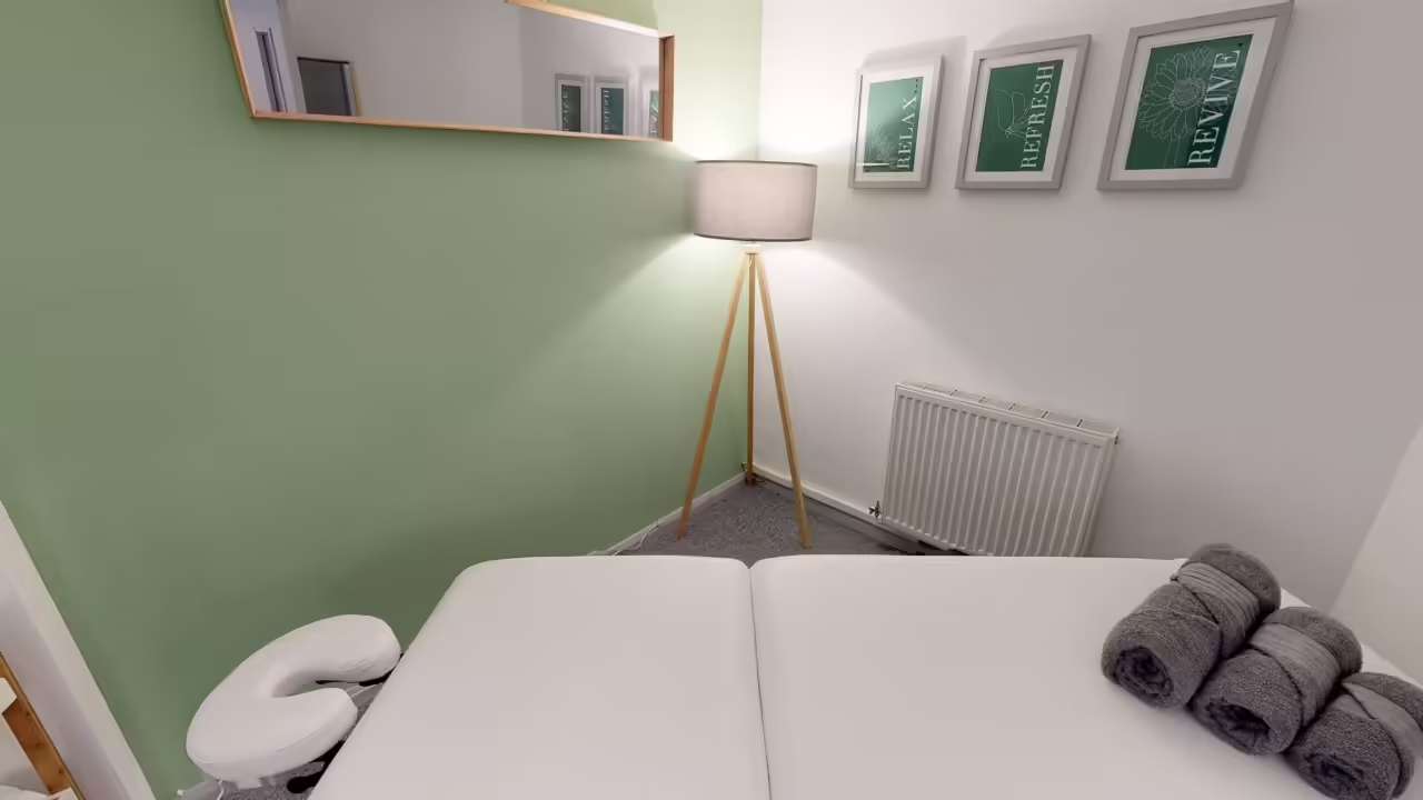 A treatment room with a white treatment couch, rolled grey towels, a tripod floor lamp and three framed prints