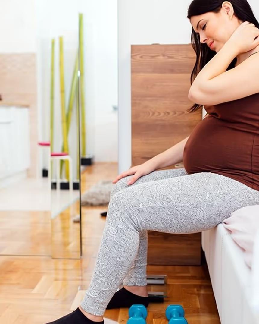 A pregnant woman sitting on the edge of a bed with one hand at the back of her neck