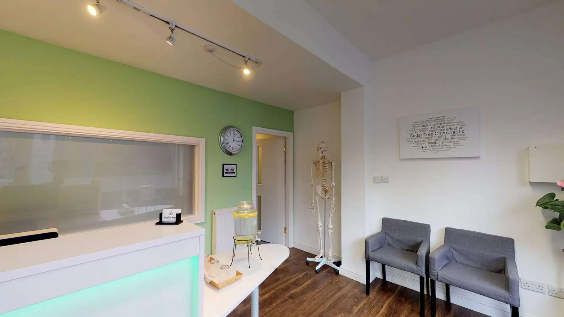A clinic reception desk and waiting area with two grey chairs, a wall clock and an anatomical skeleton model