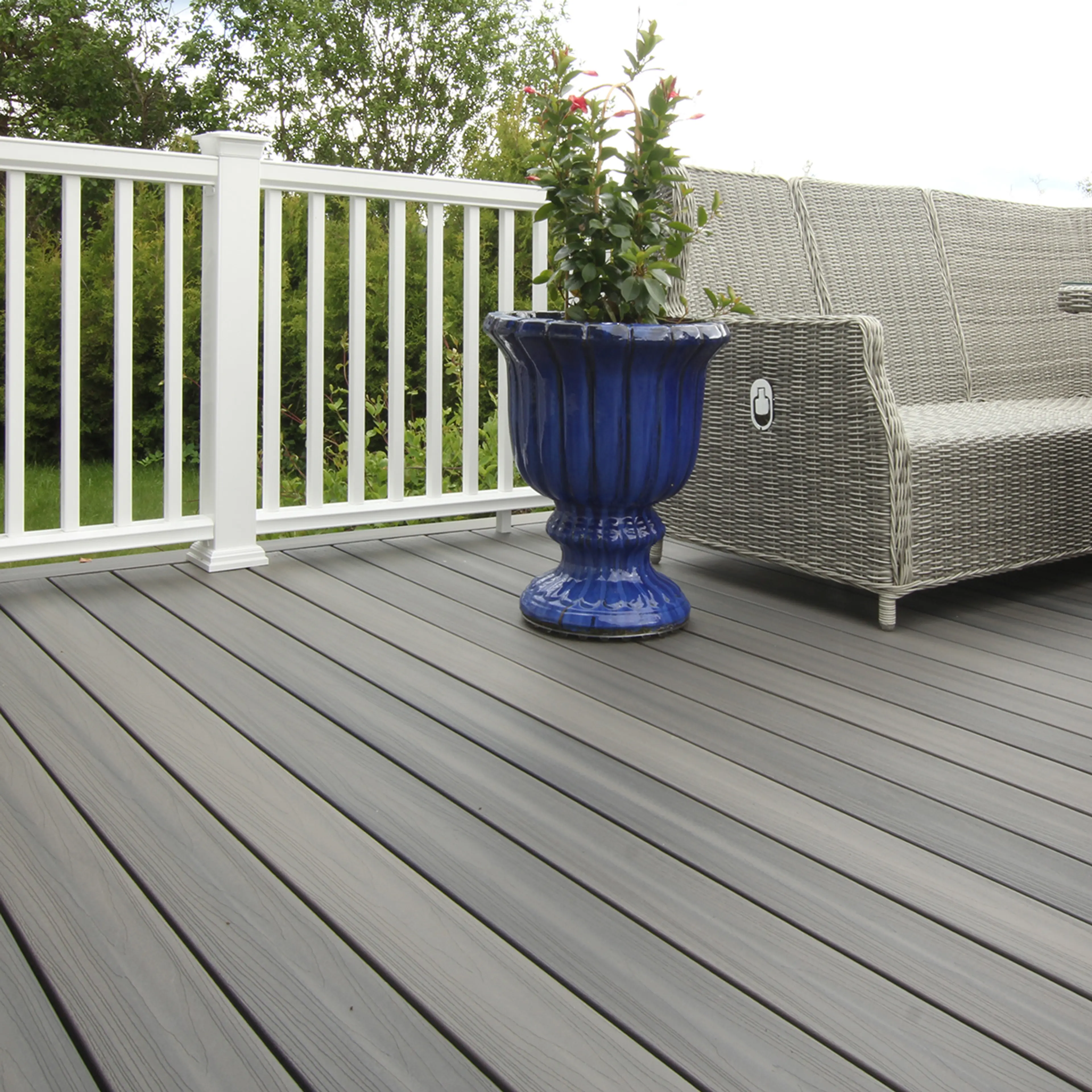 Nærbilde av terrasse med kompositt terrassegulv av typen Fiberon Sanctuary i fargen Earl Grey. Hvitt rekkverk, rottingsofa og grønn plante i blå potte