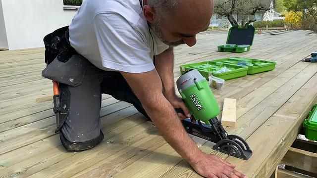 en snekker sitter på knærne på en terrasse som er under oppføring. Bruker en spikerpistol fra Essve