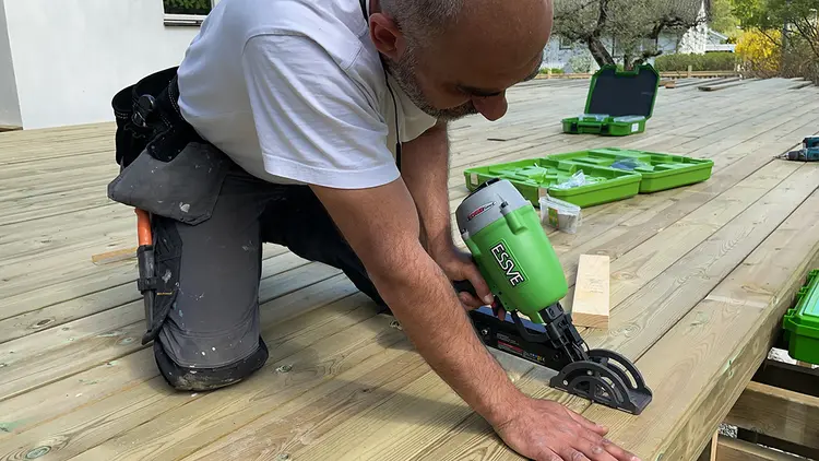 en snekker sitter på knærne på en terrasse som er under oppføring. Bruker en spikerpistol fra Essve