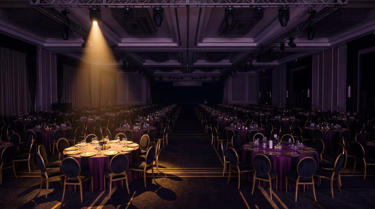 A dark, moody wide shot of an empty corporate ballroom with identical round tables set in rows — deep purple and black lighting, gold accents on the chairs, a single spotlight illuminating the predictability