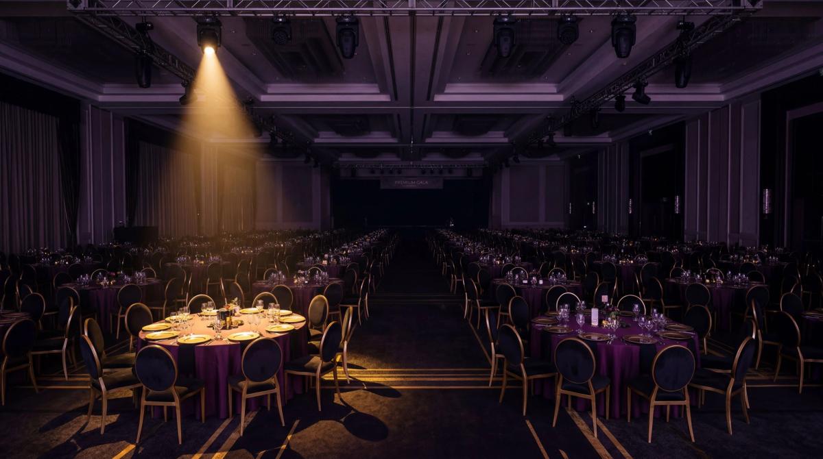 A dark, moody wide shot of an empty corporate ballroom with identical round tables set in rows — deep purple and black lighting, gold accents on the chairs, a single spotlight illuminating the predictability