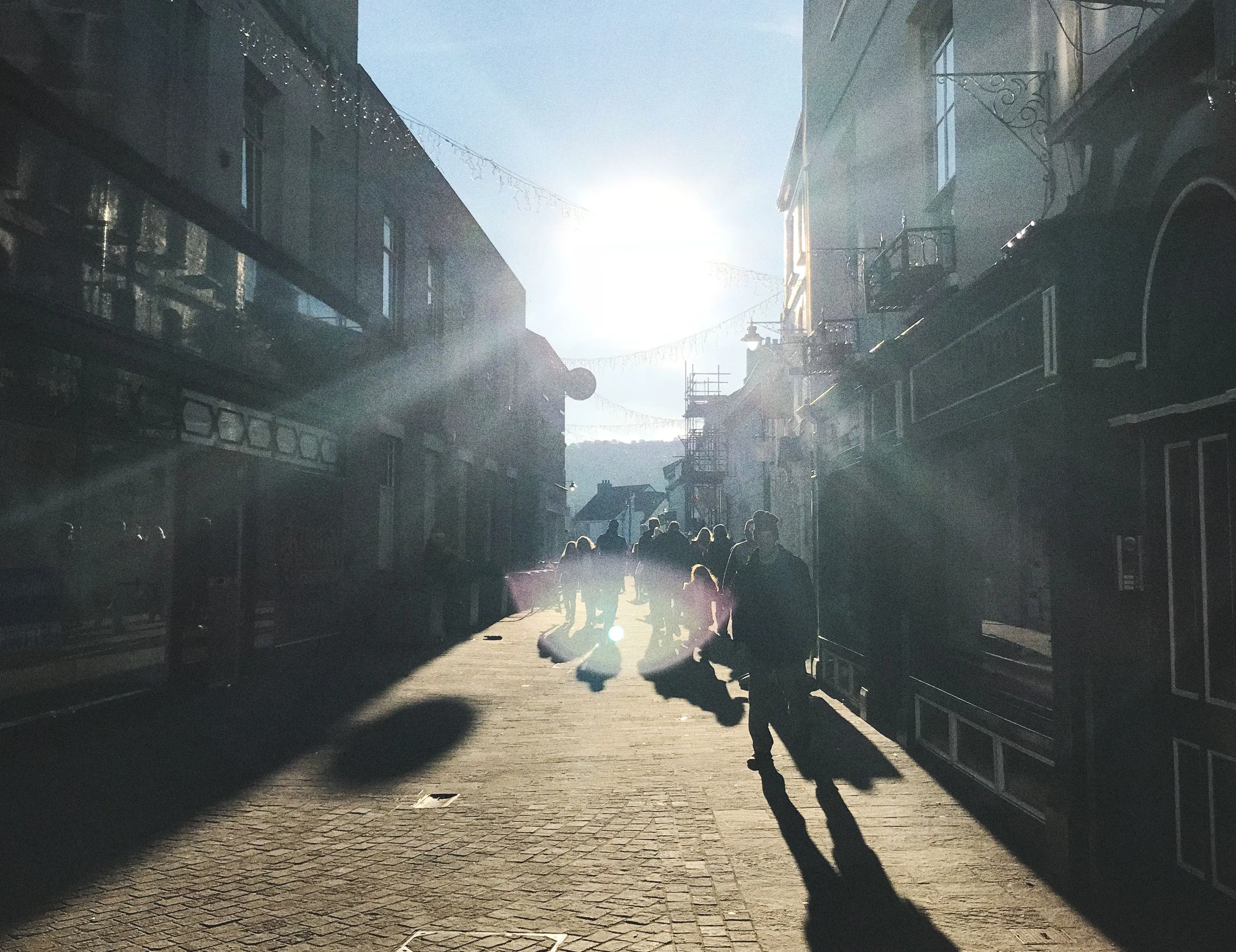 A Welsh town lit by a strong sun with people and their shadows cast long on old cobbled stones.