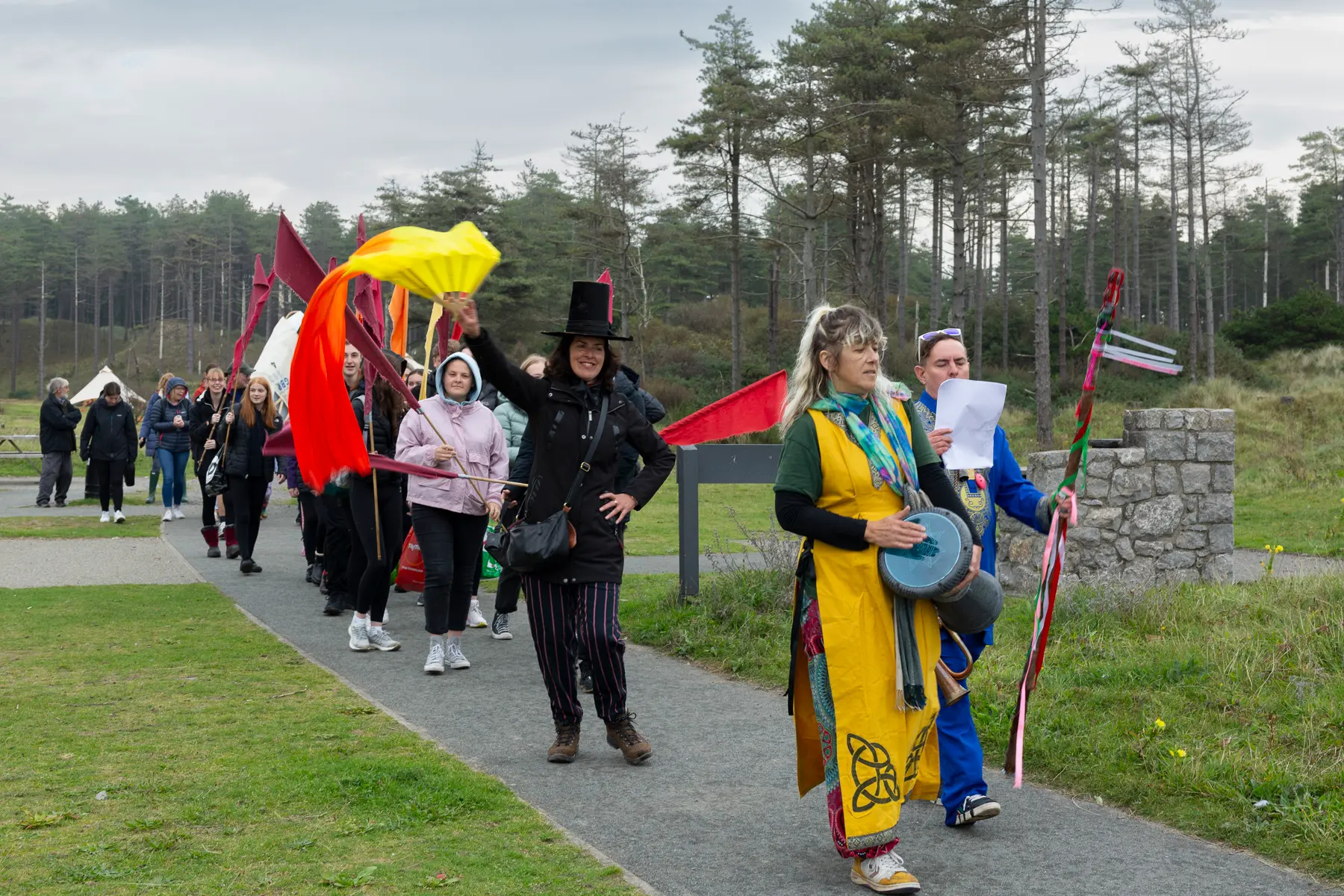 Three bards lead a procession of people waving flags along a path with a pine forest in the background.