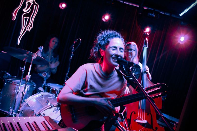 person singing on stage holding ukelele, double bass player and drummer in background