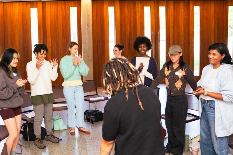 Participants standing in circle applauding. Malaika visible from behind. 