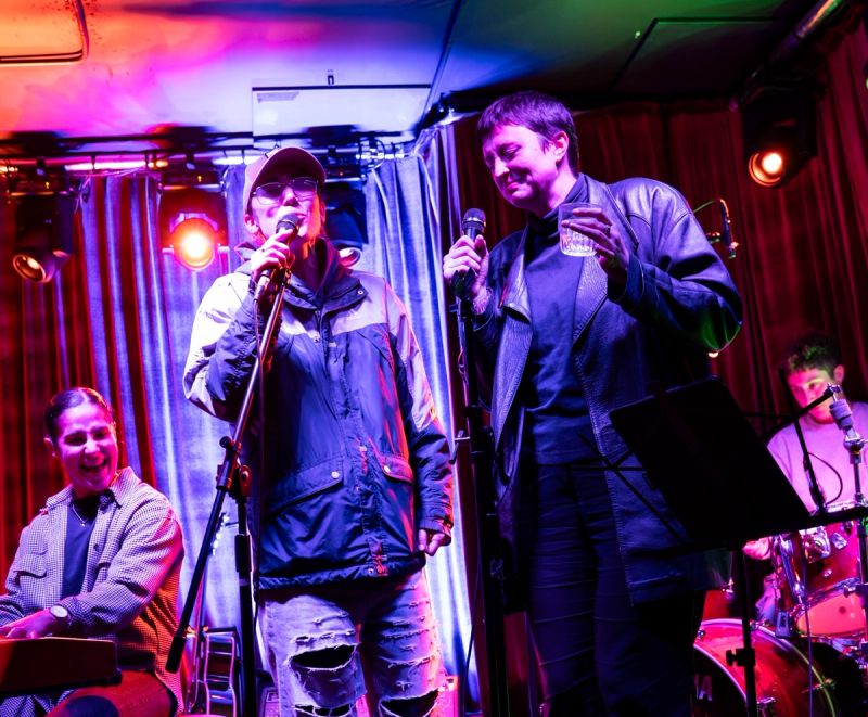 two jammers singing on stage, one with drink in hand, both smiling. Keys player behind them smiling