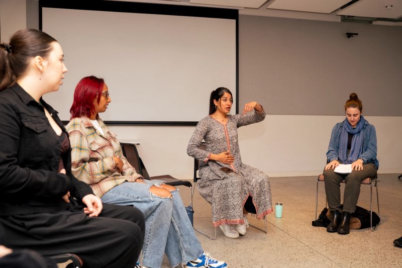 Parvyn instructing group with one hand on stomach and one raised. Everyone sitting on chairs in a circle. 3 participants visible.