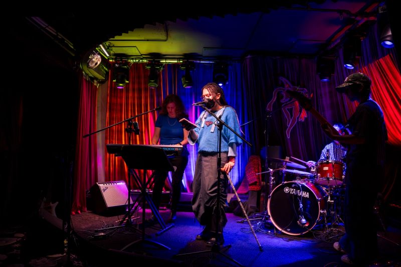 jammer on stage at microphone, reading off of phone. band surrounding them with keys, drums, and bass