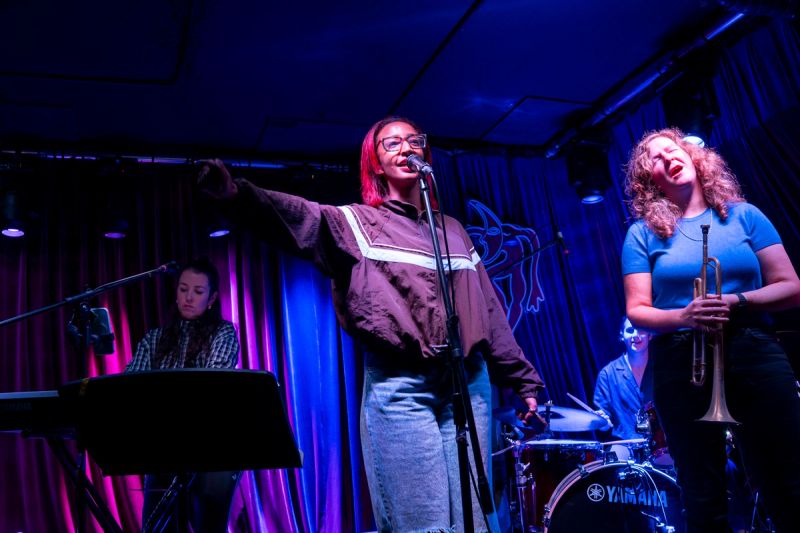 jammer on stage speaking into microphone, trumpet player to the right with eyes closed, keys player to the left playing