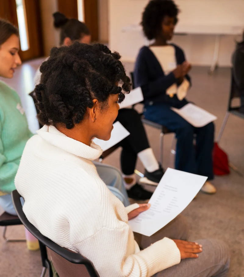 Participant sitting photographed from behind holding sheet of paper, others visible in background. All sitting on chairs in a circle.