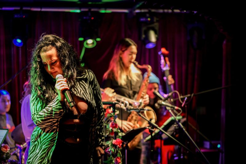 zoe K on stage looking down with mic in hand. sax player, trumpeter, bassist, and drummer visible in background