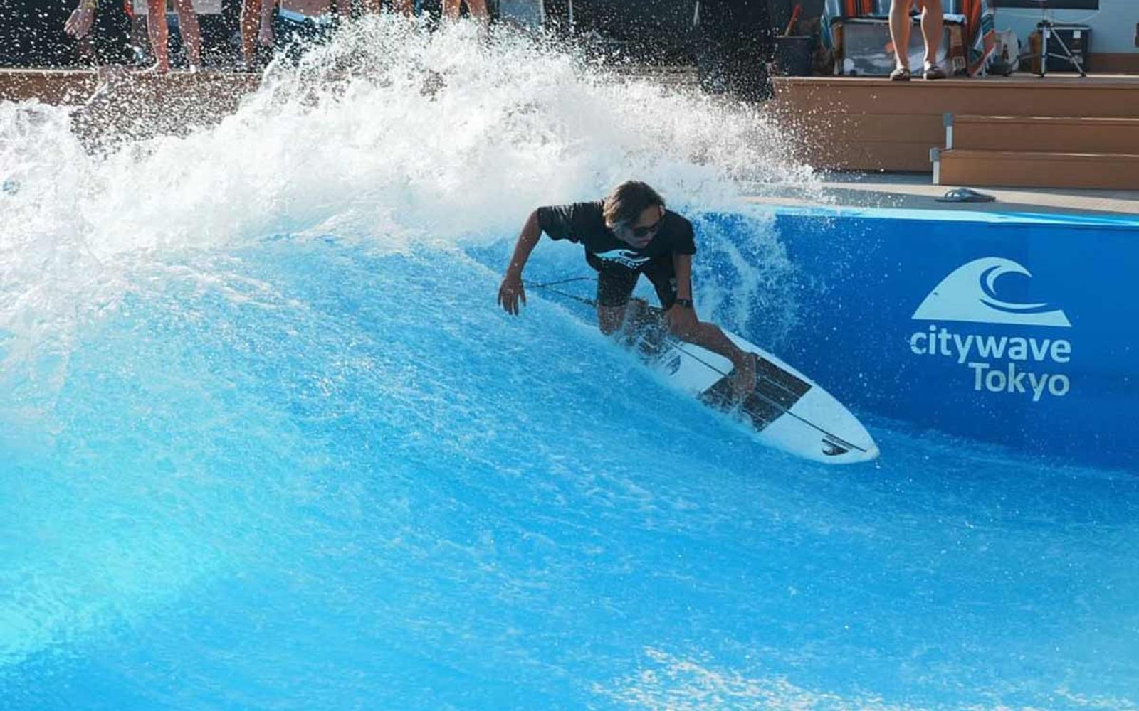 A surfer turning on the wave at Citywave Tokyo, with the pool's branded wall behind.