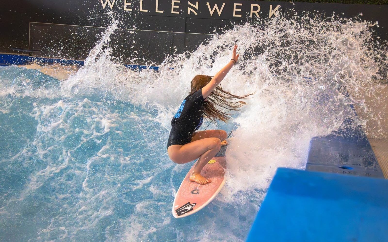 A surfer throws spray on Wellenwerk's indoor wave in Berlin, under the venue's sign.