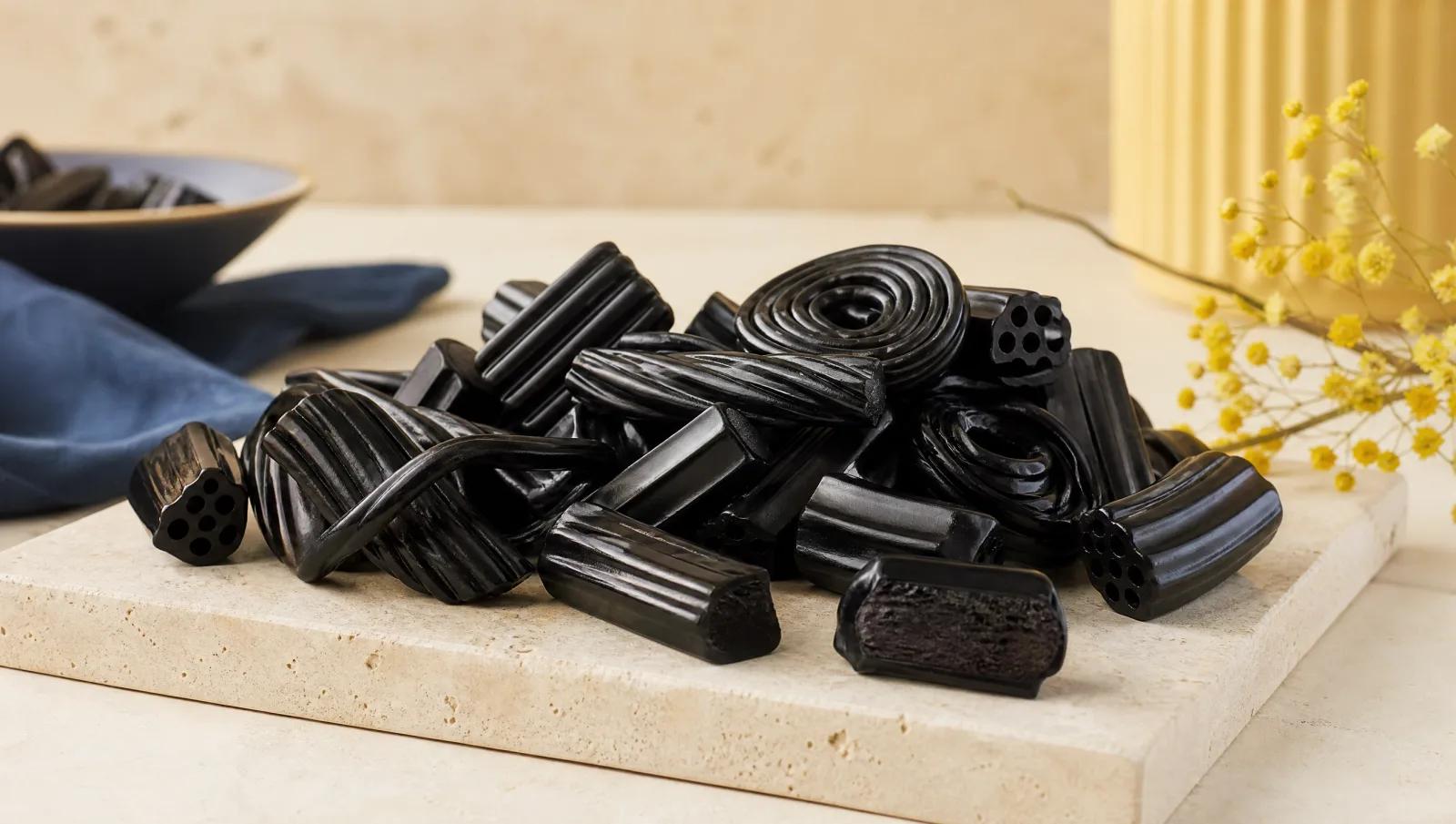Glossy black licorice pieces in varied shapes heaped on cream stone, one broken to show the interior.