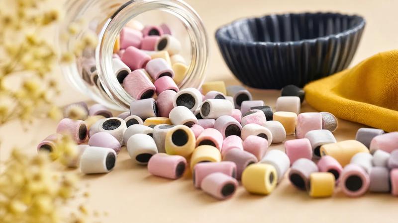 Pastel sugar-shelled licorice pellets spilling from a small glass jar, macro texture.