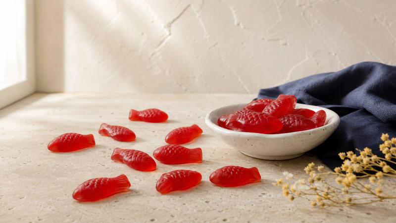 Red fish-shaped gummy candies scattered on cream stone, with a few in a small white dish.