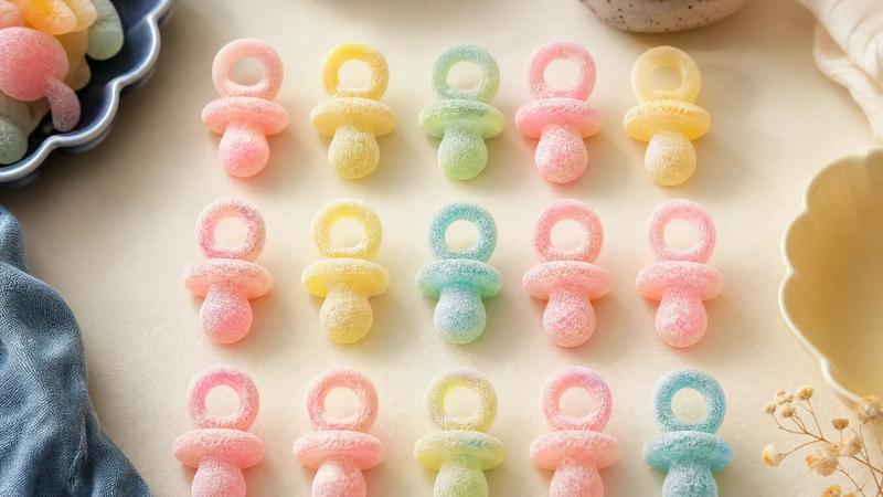 Sugar-dusted pacifier-shaped sour sweets in pastel colours, overhead in rows.