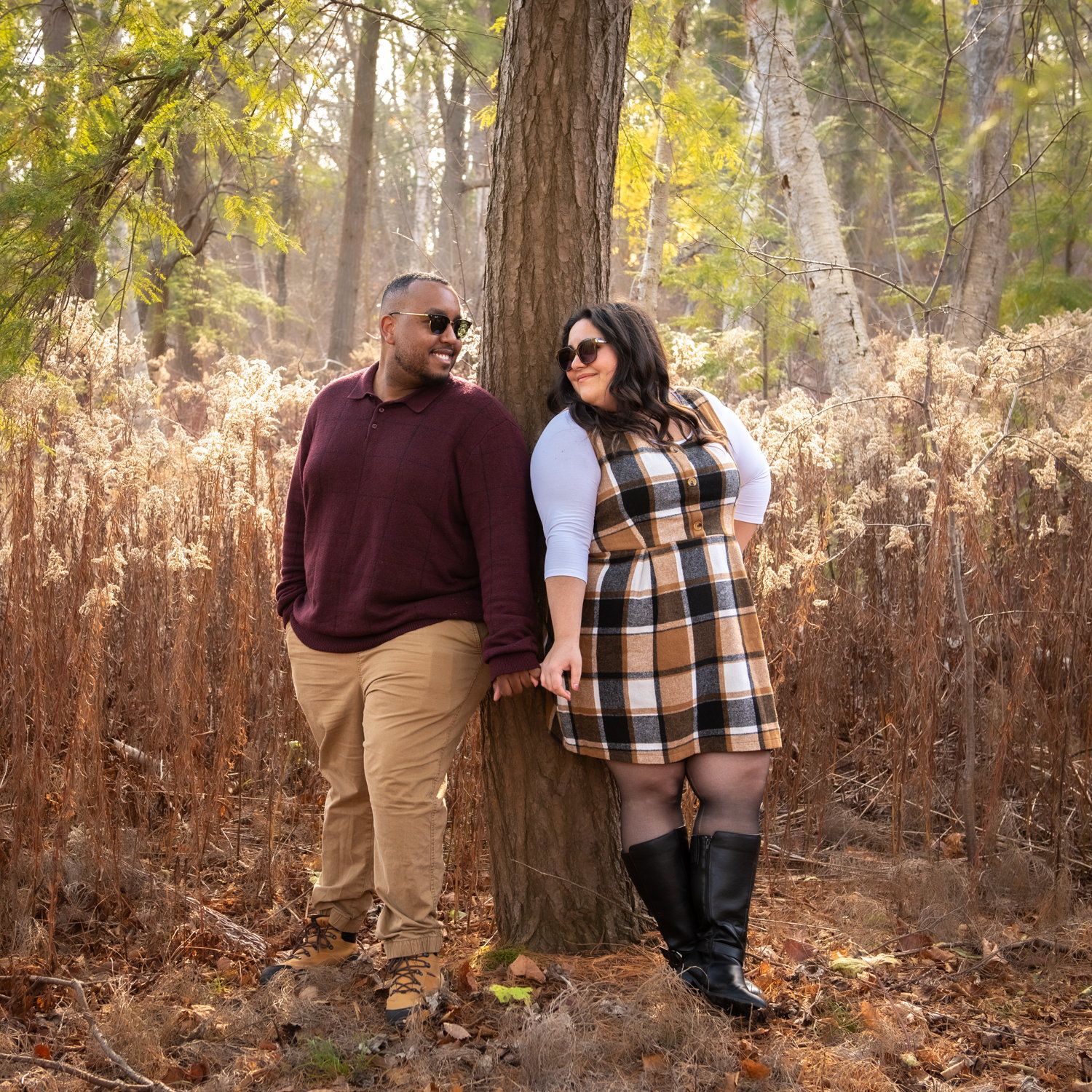A couple in autumn layers standing against a broad tree trunk in dry woodland