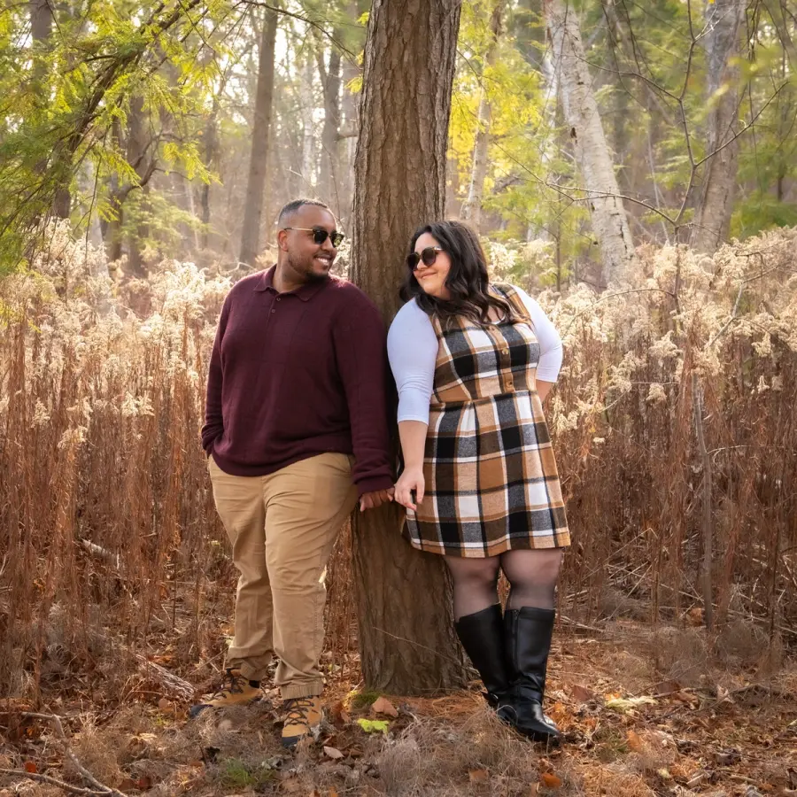 A couple in autumn layers standing against a broad tree trunk in dry woodland