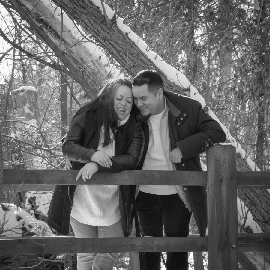 Black-and-white photo of a couple laughing together on a snowy wooden fence