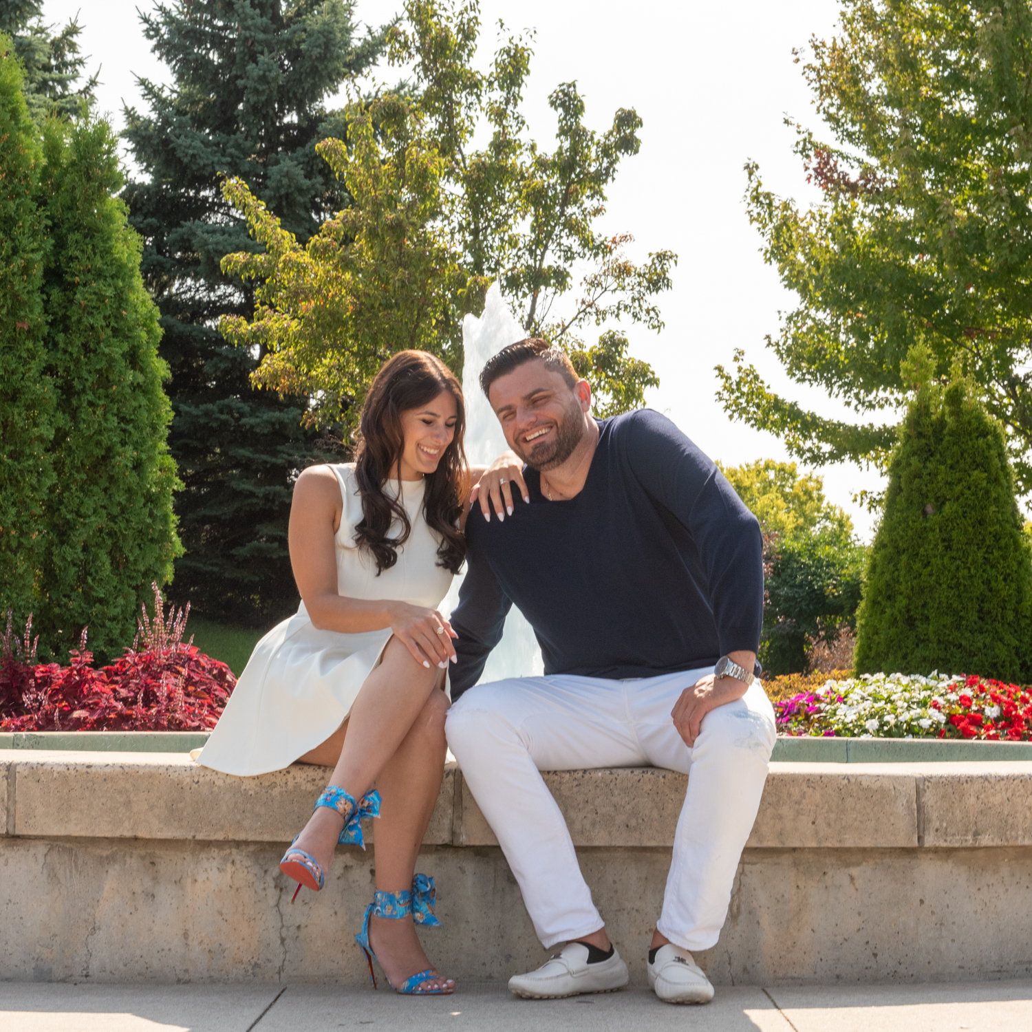 A couple sitting on the stone rim of a garden fountain among red and pink flowers
