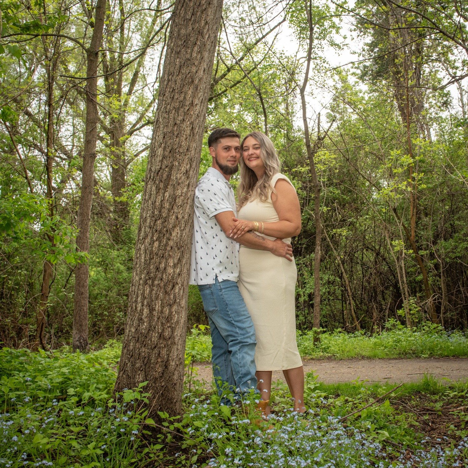 A couple leaning against a tall pine on a woodland path in late spring