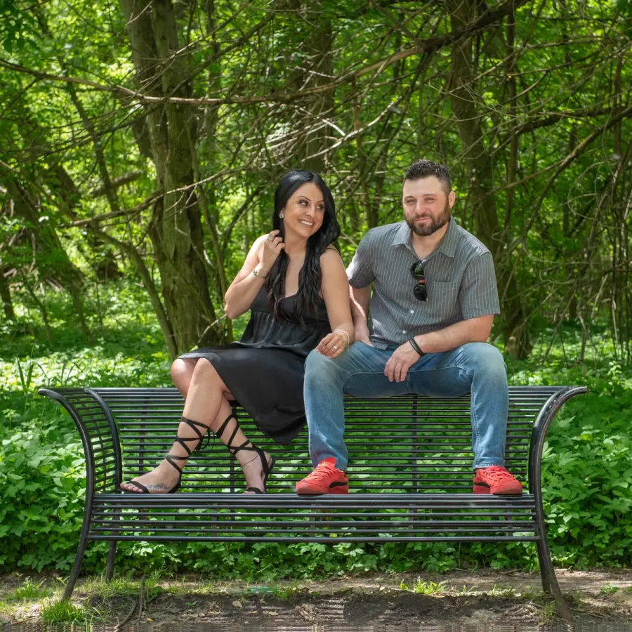 A couple sitting at opposite ends of a park bench under heavy green trees