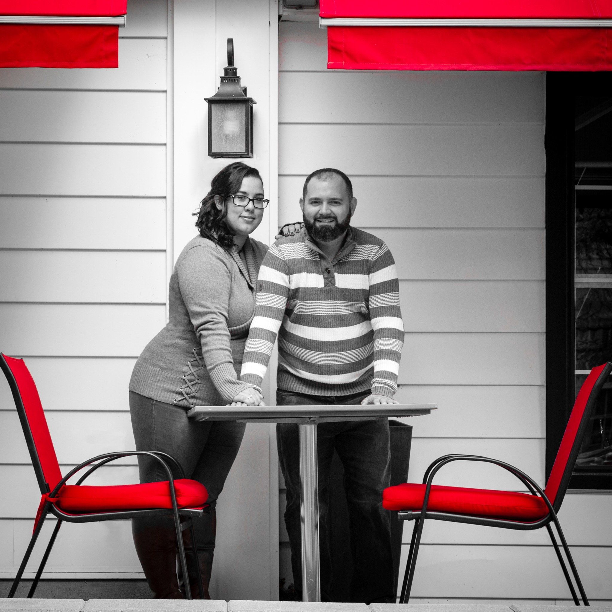 Black-and-white photo of a couple at a cafe table, with the red chairs and awning left in colour