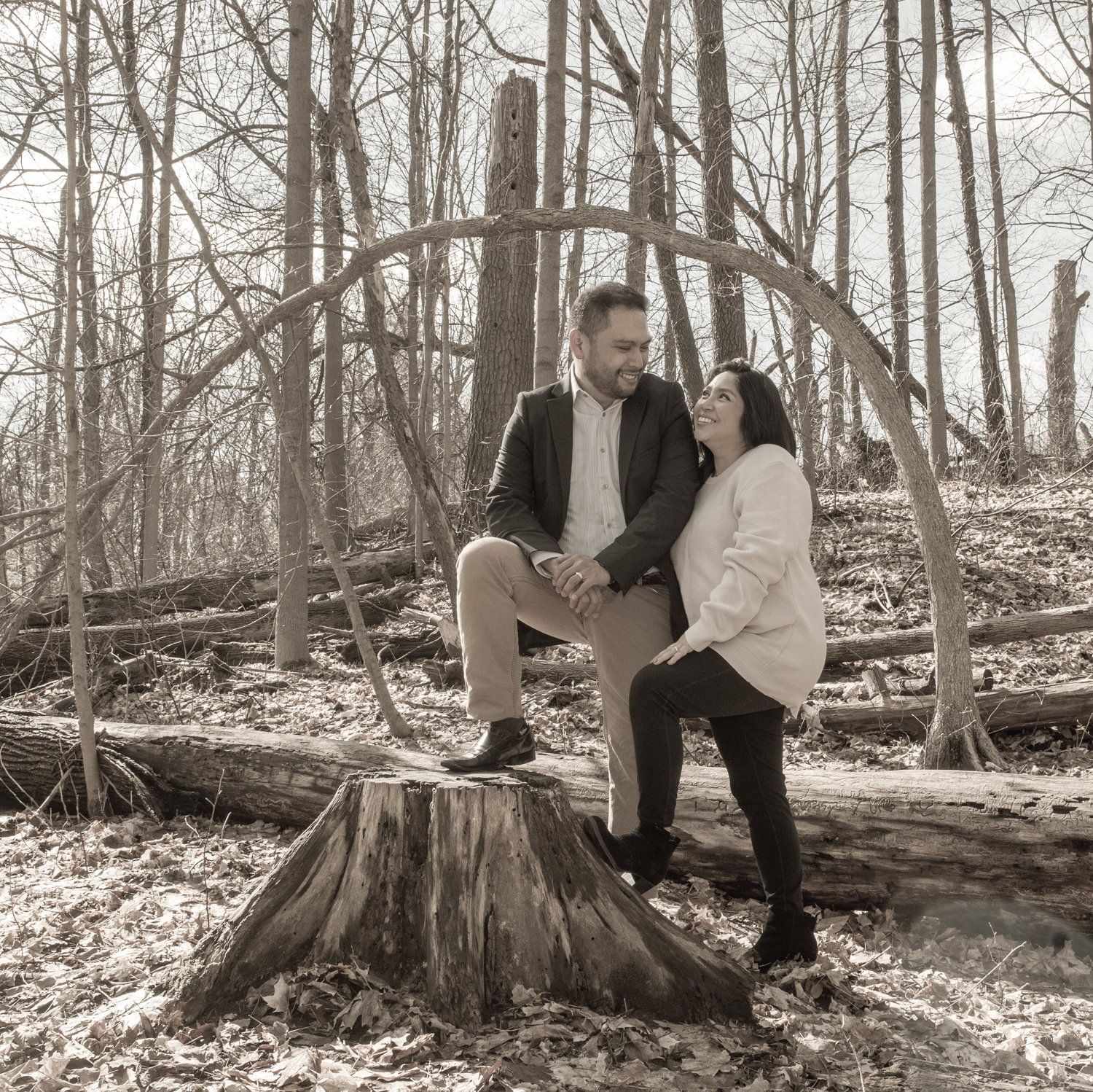 Black-and-white photo of a couple perched on a tree stump in a bare winter wood