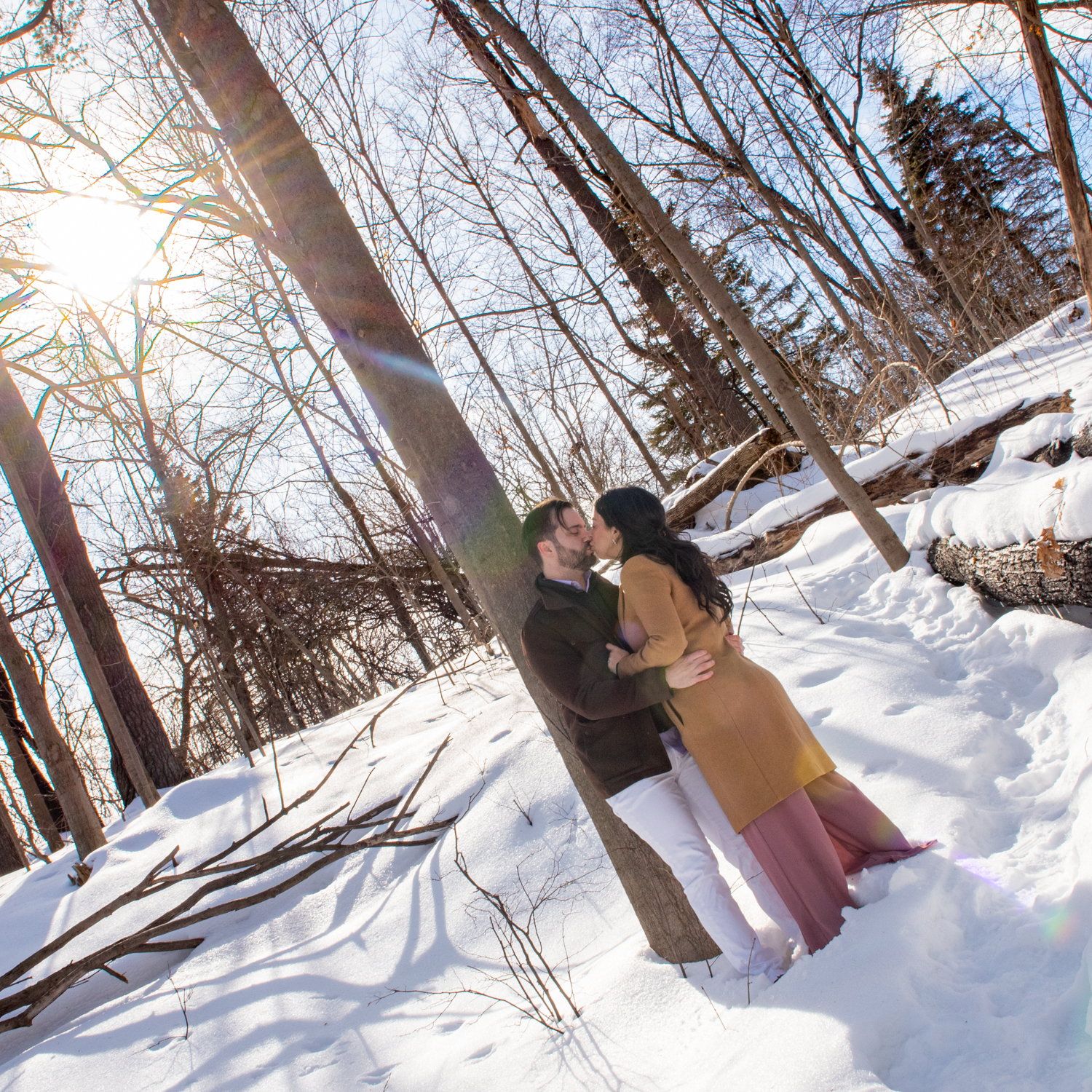 Kisa & Larry embracing against a tree on a snowy slope, sun flaring through