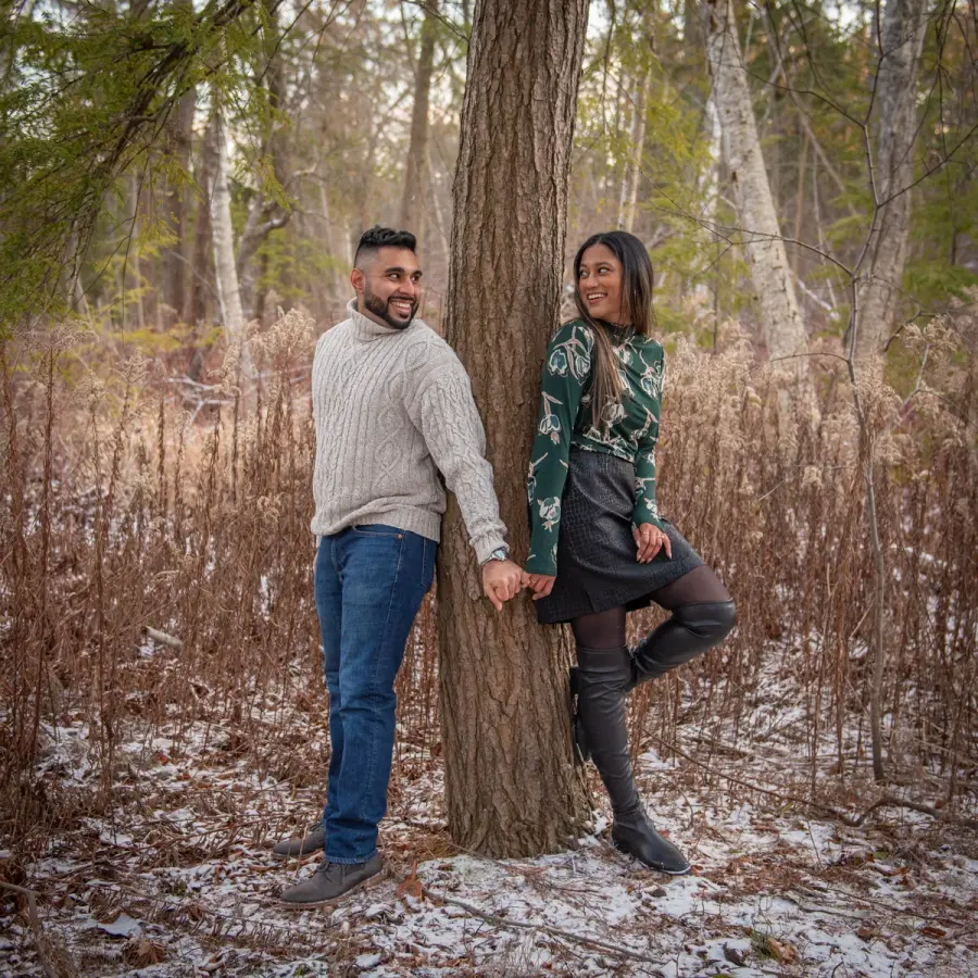 A couple holding hands on either side of a tree trunk in a snow-dusted wood