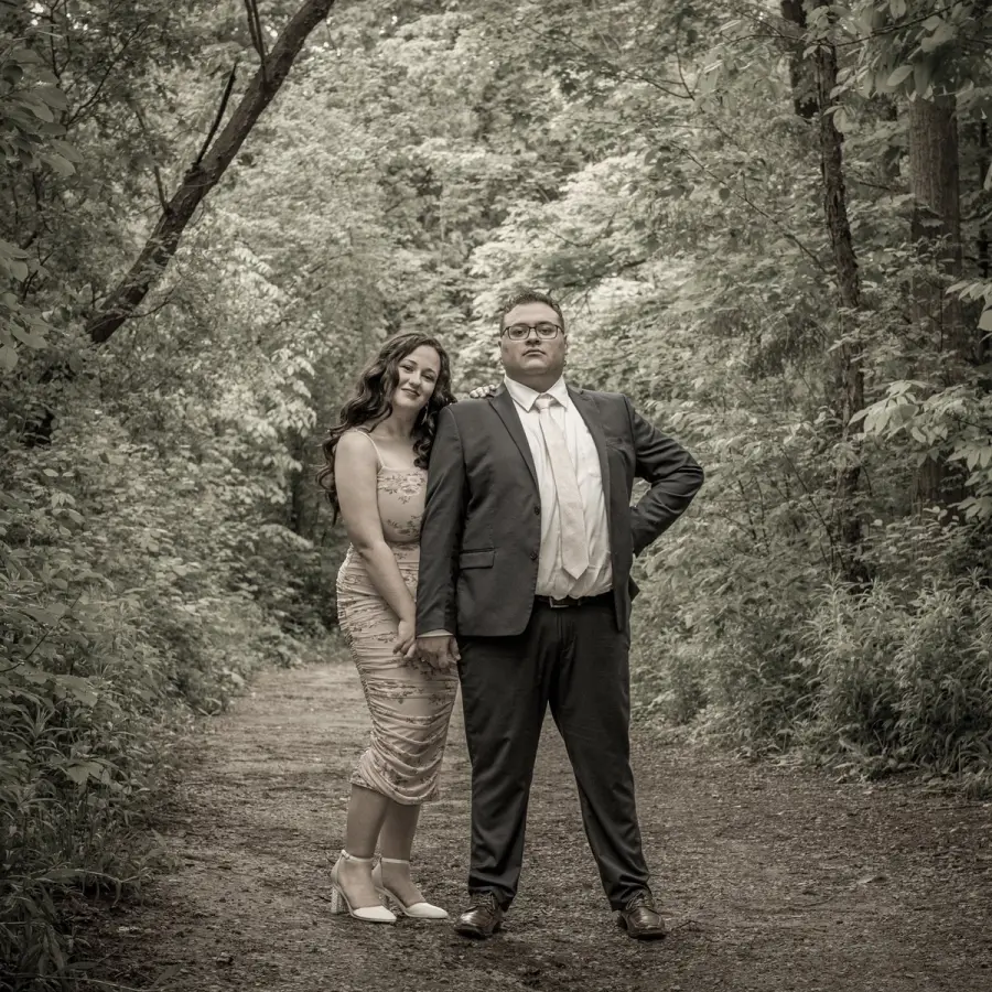 Black-and-white photo of a couple in formal dress standing on a wooded park path
