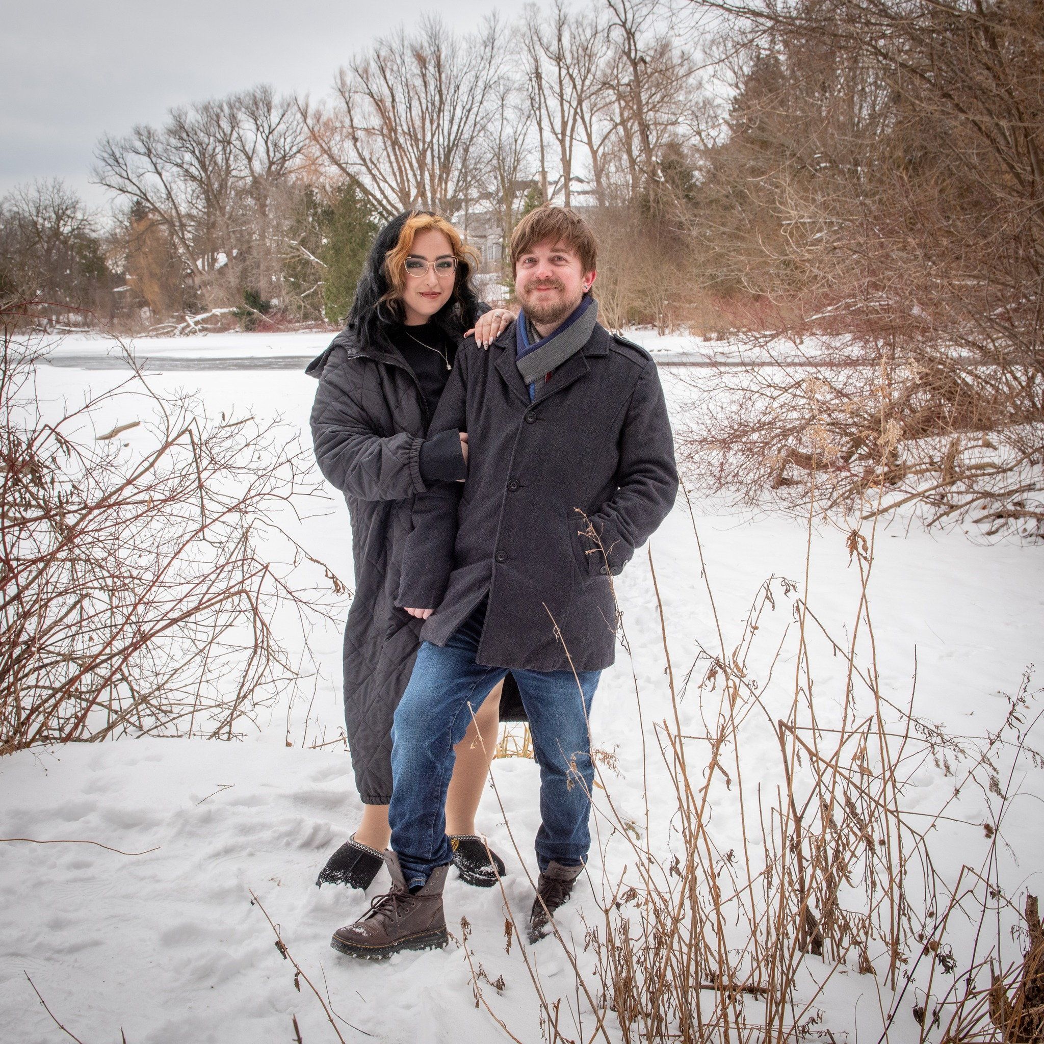 A couple in winter coats standing in snow beside dry golden grasses
