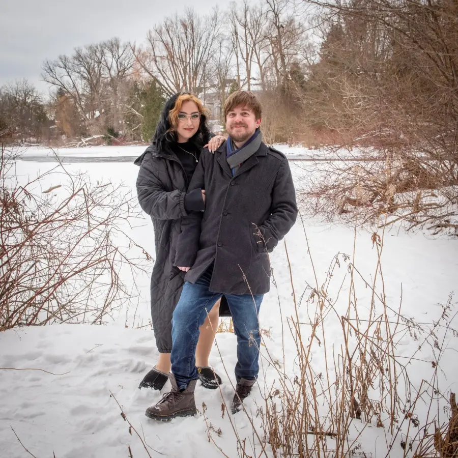 A couple in winter coats standing in snow beside dry golden grasses