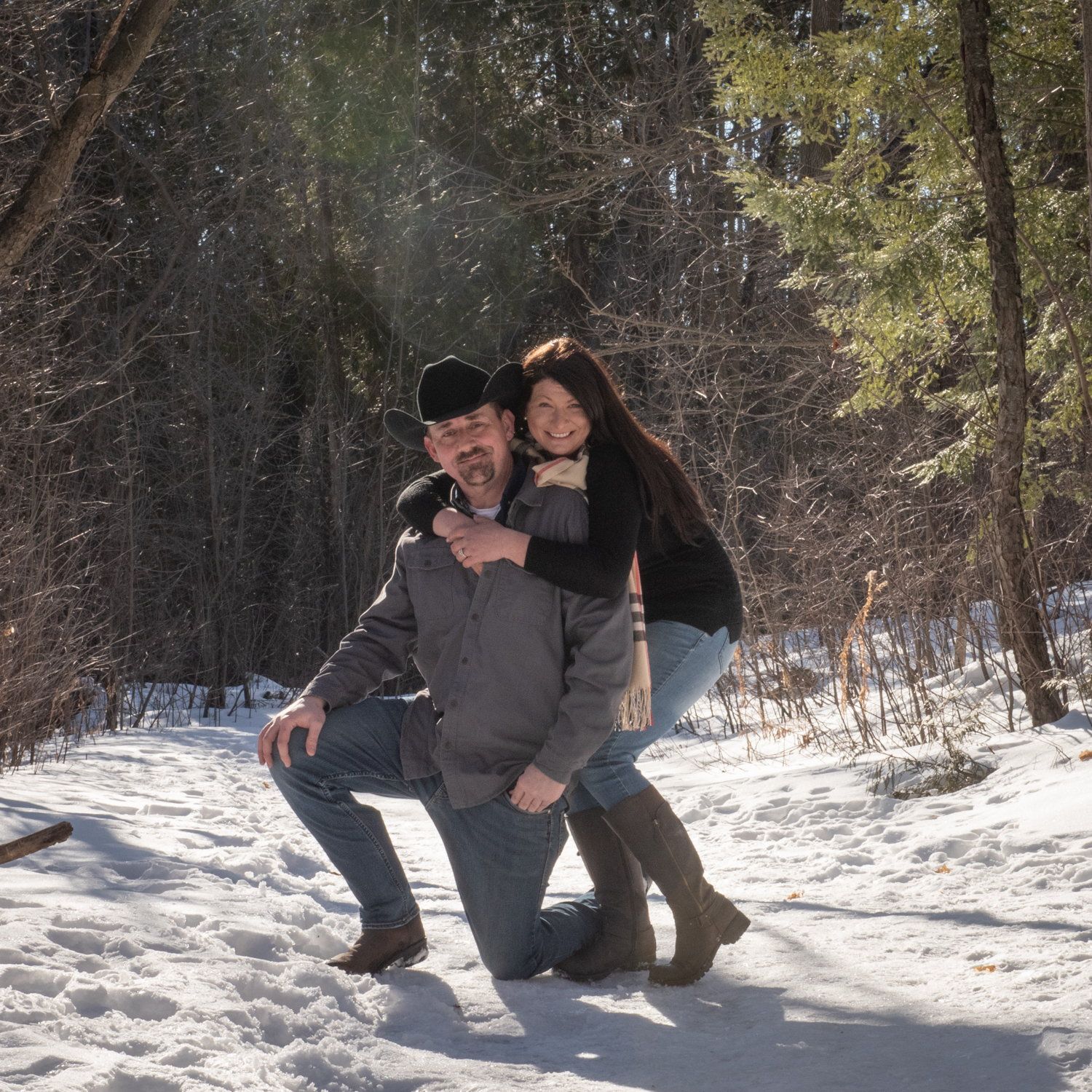 A man kneeling in the snow with his partner leaning on his shoulder, pines behind them