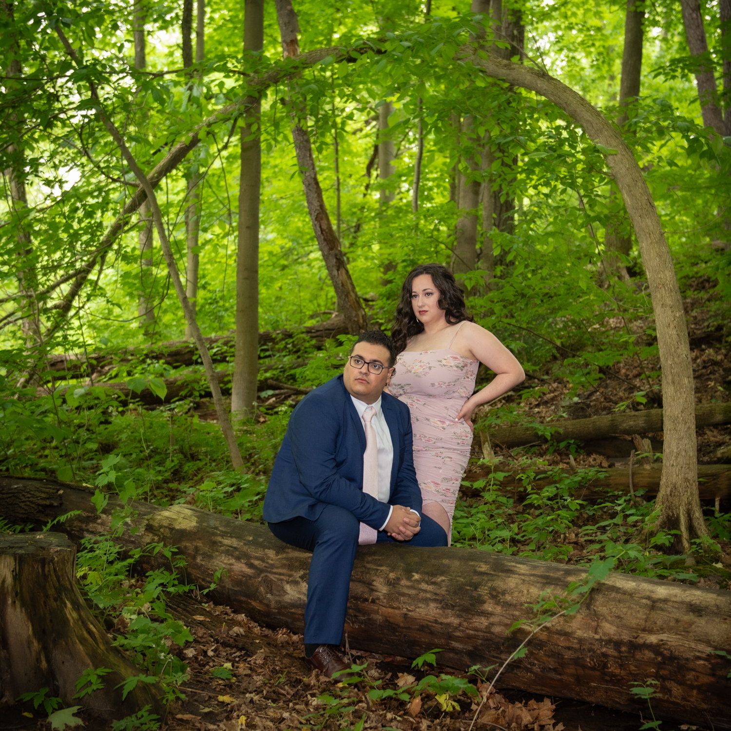 A couple in a navy suit and blush gown resting on a mossy fallen log in a summer forest