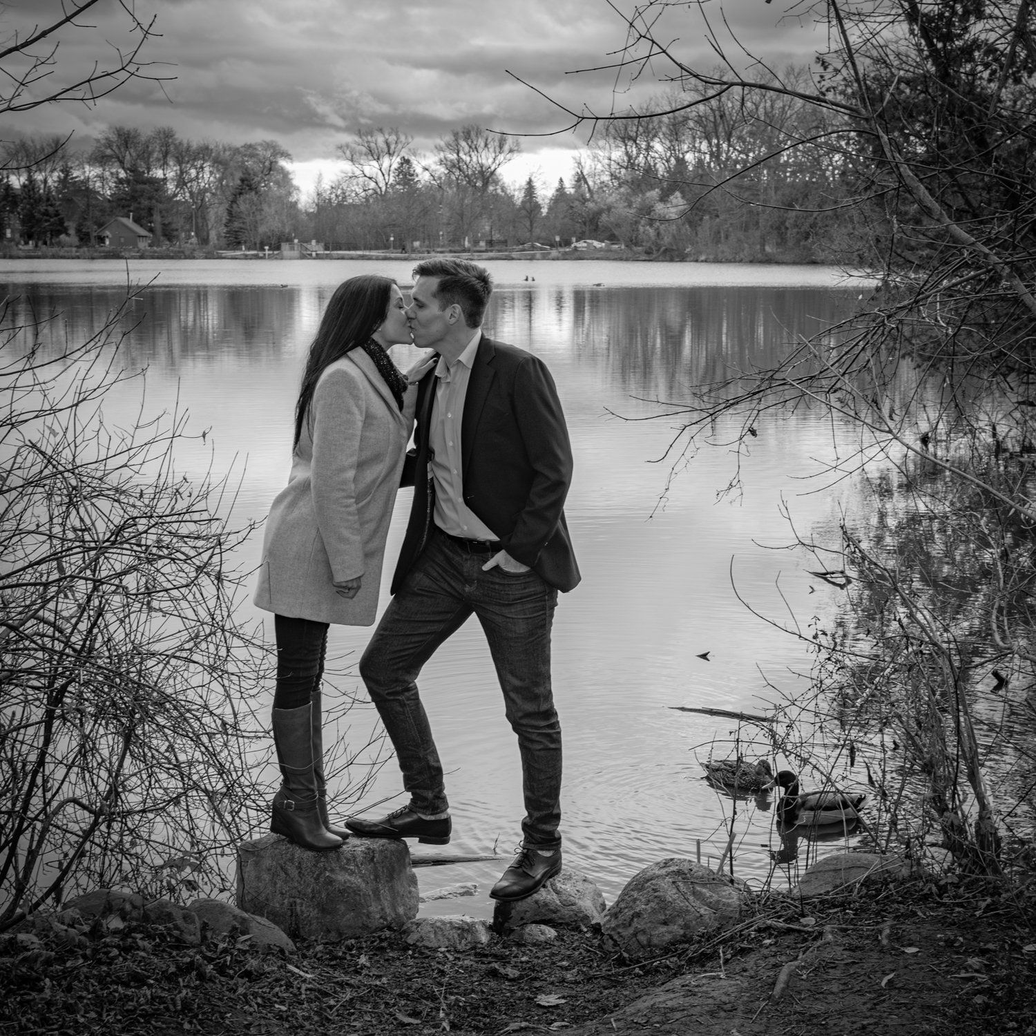 Black-and-white photo of a couple kissing on rocks at the edge of a lake, ducks on the water