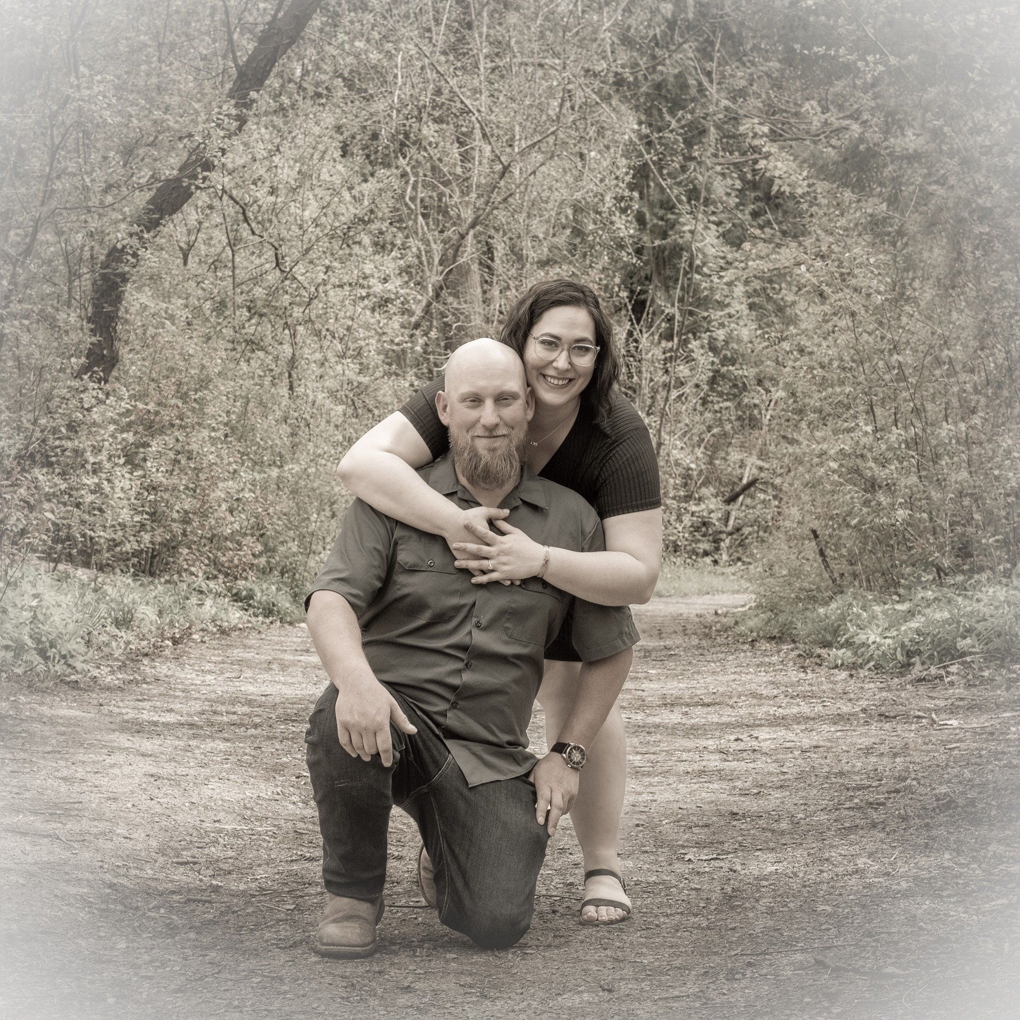Black-and-white photo of a man kneeling on a park path while his partner leans over his shoulder
