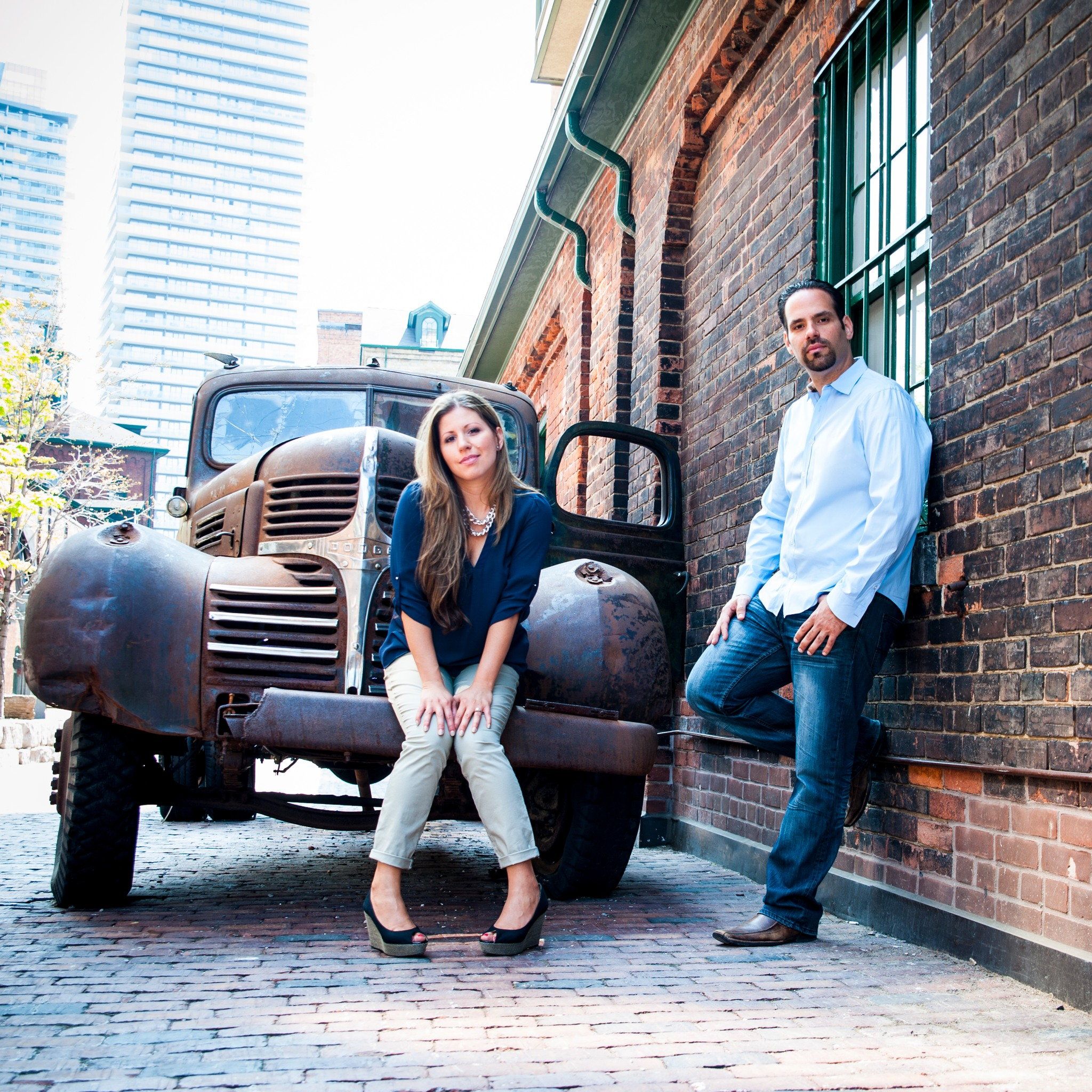 A couple leaning on a vintage pickup truck beside a brick warehouse downtown